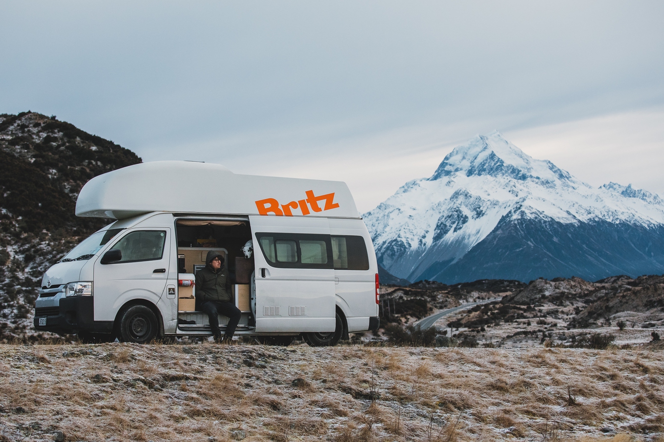 A man on a roadtrip holiday sits in a campervan in front of a mountain