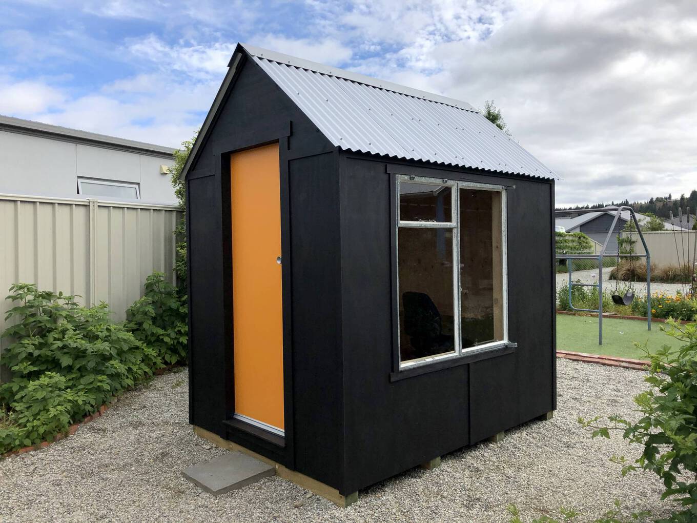 A photo of the finished Writer's Studio - it has a corrugated iron roof, orange door and is painted black with a big window on the side